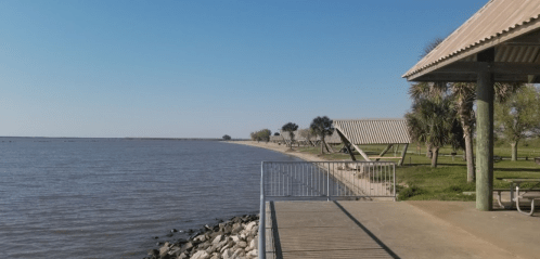 A serene waterfront view with a pathway, grassy area, and shaded pavilions under a clear blue sky.