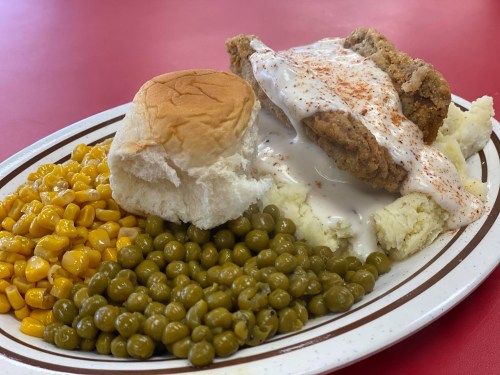 A plate of fried chicken with gravy, mashed potatoes, corn, green peas, and a soft roll on a red table.