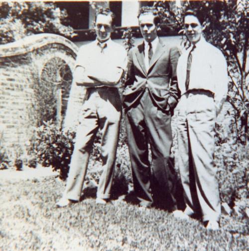 Three men in suits stand together outdoors, with greenery and a brick wall in the background.