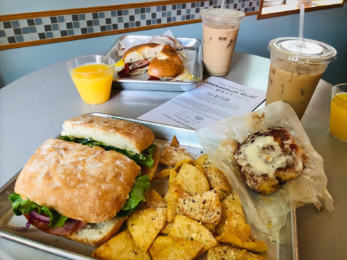 A table with a sandwich, seasoned potato chips, a cinnamon roll, and iced drinks, with a menu in the background.