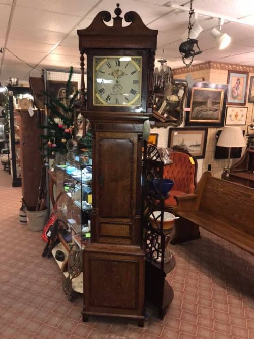 A tall, vintage wooden grandfather clock with a decorative face, surrounded by various antique items in a shop.