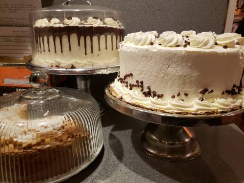 Three cakes on display: a chocolate-drizzled cake, a white frosted cake with swirls, and a layered cake under a glass dome.