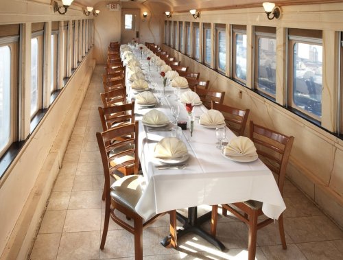 A long dining table set with white tablecloths, folded napkins, and roses, inside a train car with large windows.