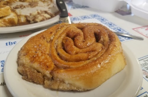 A freshly baked cinnamon roll on a white plate, glistening with a sugary glaze.
