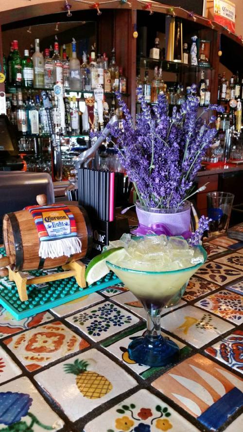 A colorful bar scene featuring a margarita glass, a small barrel, and a vase of lavender flowers on a tiled counter.