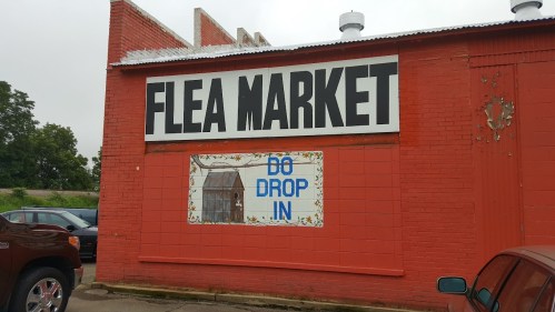 A red brick building with a large sign reading "Flea Market" and a smaller sign saying "DO DROP IN."