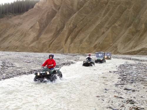 Three ATV riders navigate a rocky riverbed, splashing through water with steep, rugged cliffs in the background.