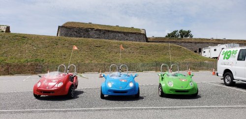 Three colorful go-karts (red, blue, green) parked in a lot with a grassy hill and a van in the background.