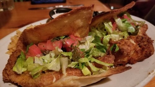 Two crispy taco shells filled with lettuce, tomatoes, and fried fish, served with rice on a white plate.