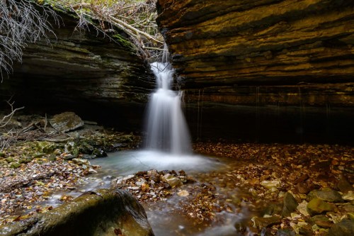 A serene waterfall cascades over rocks into a tranquil pool, surrounded by mossy cliffs and fallen leaves.