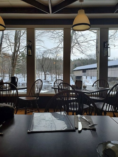 A cozy restaurant interior with wooden chairs, a table set for dining, and a snowy landscape visible through large windows.