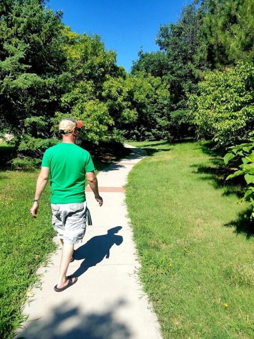 A person in a green shirt walks along a sunny path surrounded by trees and greenery.