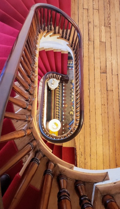 A spiral staircase with wooden railings, red carpet, and a patterned floor, viewed from above.