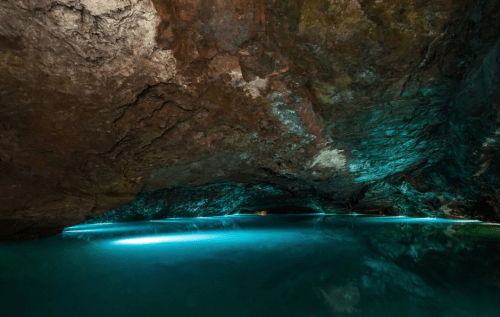 A serene underground cave with a calm, illuminated water surface reflecting the rocky ceiling above.