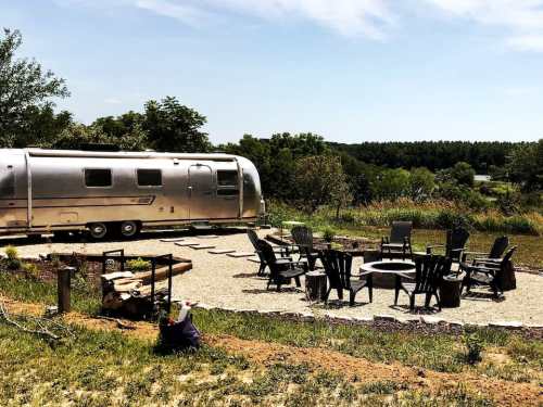 A silver Airstream trailer beside a gravel patio with black chairs and a fire pit, surrounded by greenery and trees.