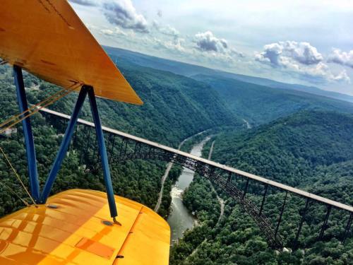 Aerial view of a yellow biplane flying over a lush green valley and a bridge spanning a river below.