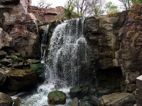 A cascading waterfall flows over rocky terrain, surrounded by greenery and natural stone formations.