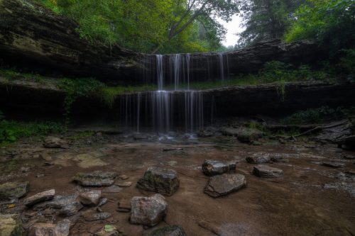 A serene waterfall cascades over rocky ledges, surrounded by lush greenery and a tranquil stream below.