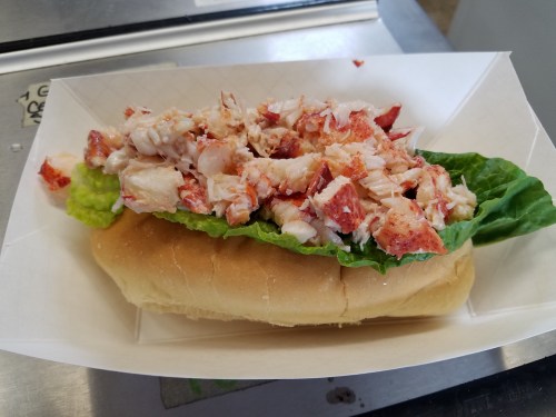 A lobster roll topped with fresh lobster meat and lettuce on a soft bun, served in a paper tray.
