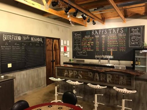 Interior of a bar with a chalkboard menu, wooden counter, and seating, featuring beer options and prices.