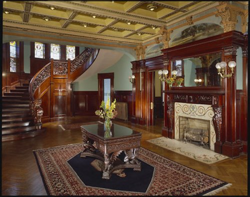Elegant interior of a grand foyer featuring a staircase, ornate woodwork, stained glass, and a decorative fireplace.