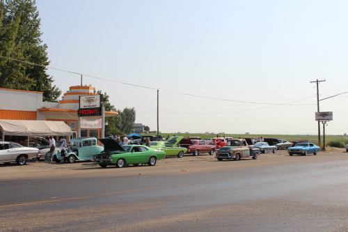 A classic car show outside a diner, featuring colorful vintage cars parked along a rural road.