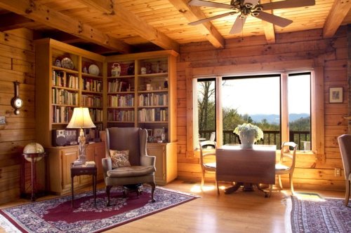 Cozy wooden living room with a bookshelf, armchair, dining table, and large window showcasing a mountain view.