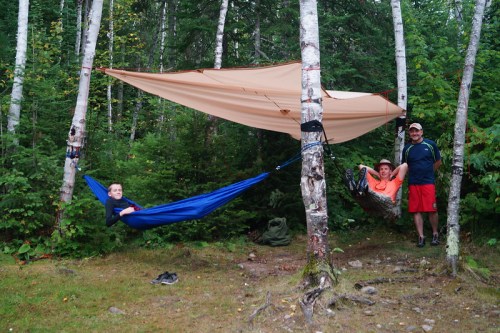A family enjoys a camping spot with two hammocks set between trees, surrounded by lush greenery.
