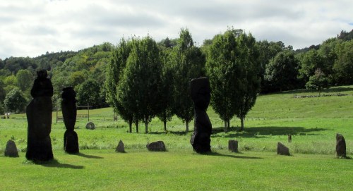 A grassy field with tall, dark stone sculptures and trees in the background under a cloudy sky.