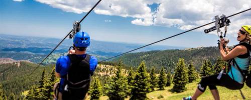 Two people ziplining over a scenic mountain landscape with trees and blue skies in the background.