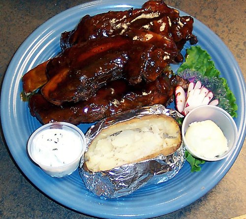 A plate of barbecue ribs with a baked potato, sour cream, and a side of lettuce and radishes.