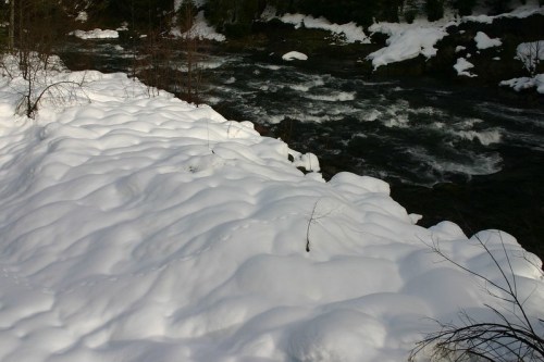 A snowy riverbank with soft, undulating snow and a flowing river in the background.