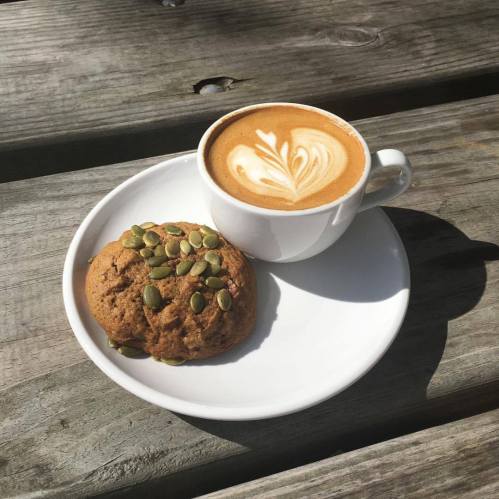 A cup of latte art coffee beside a cookie topped with pumpkin seeds on a white plate, resting on a wooden table.