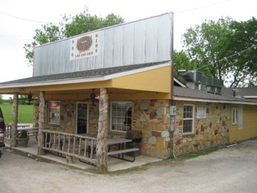 A rustic restaurant with a stone exterior and a sign reading "Ribs" on the roof, surrounded by greenery.