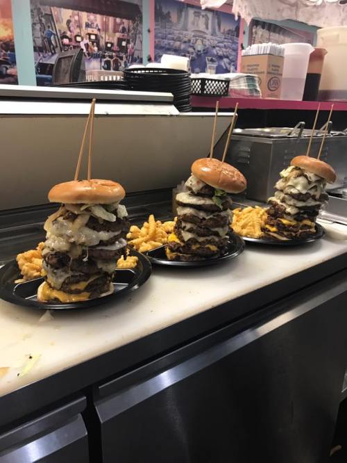 Three towering burgers with multiple patties, topped with onions and pickles, served with fries on a counter.
