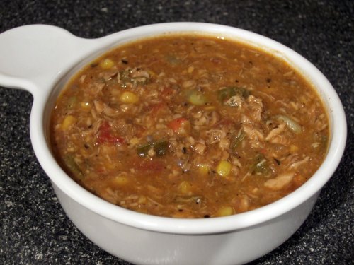 A bowl of hearty chicken soup with vegetables and rice, served in a white dish on a dark countertop.