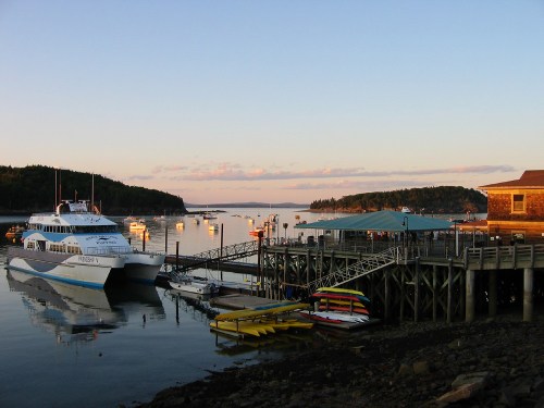 A serene harbor at sunset with boats docked, a wooden pier, and colorful kayaks on the shore.