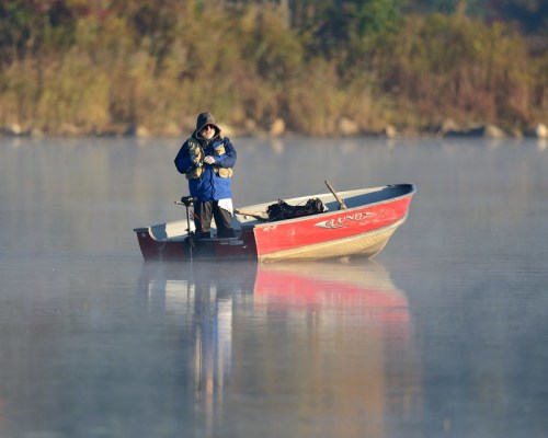 A person in a blue jacket stands in a small red boat on a misty lake, surrounded by autumn foliage.