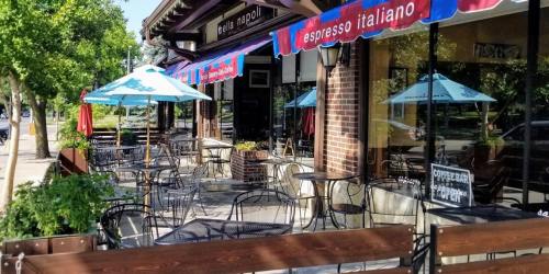 Outdoor seating area of an Italian café with blue umbrellas and a brick facade, surrounded by greenery.