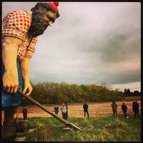 A giant statue of a lumberjack looms over a group of people standing in a field under a cloudy sky.
