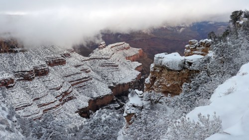 A snowy landscape of the Grand Canyon, with layered rock formations and clouds hovering above the canyon walls.