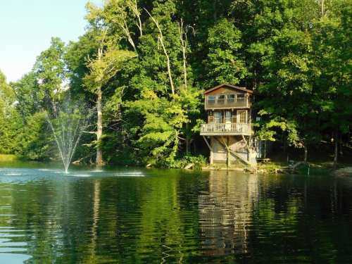 A serene lake scene featuring a wooden cabin surrounded by lush green trees and a fountain in the water.