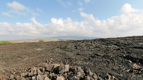 A vast, rocky landscape with black lava formations under a cloudy sky and distant hills in the background.