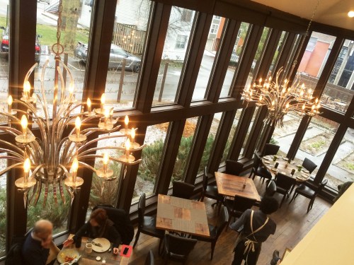 A modern restaurant interior with large windows, chandeliers, and diners at tables.