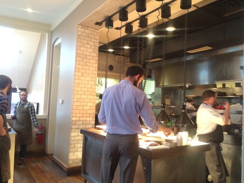 A busy restaurant kitchen with chefs preparing food and staff working at a counter. Bright, modern interior design.