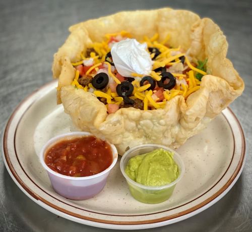 A taco salad in a crispy shell, topped with cheese, olives, tomatoes, and sour cream, served with salsa and guacamole.