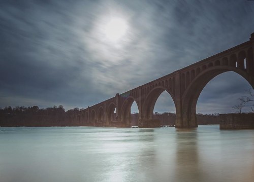A long bridge with arches spans a calm river under a bright, cloudy sky, illuminated by the sun.