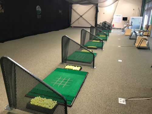 Indoor golf practice area with multiple hitting bays, each equipped with green mats and baskets of golf balls.