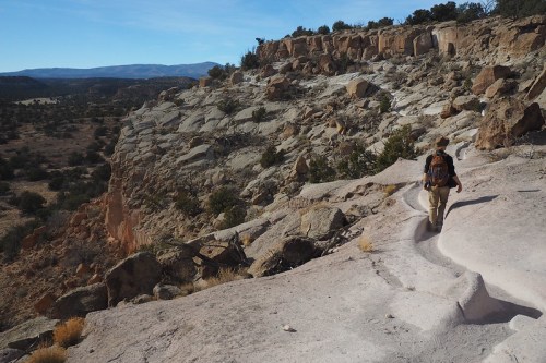 A hiker walks along a rocky trail on a cliffside, with expansive desert landscape and mountains in the background.