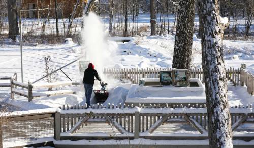 A person using a snowblower to clear snow from a driveway in a winter landscape. Trees and a fence are visible.